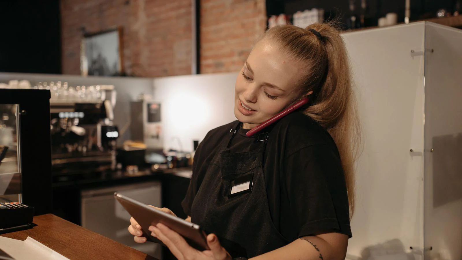Atendente de uma cafeteria conversa ao telefone e consulta um tablet atrás do balcão, em uma rotina de pedidos e atendimento digital.